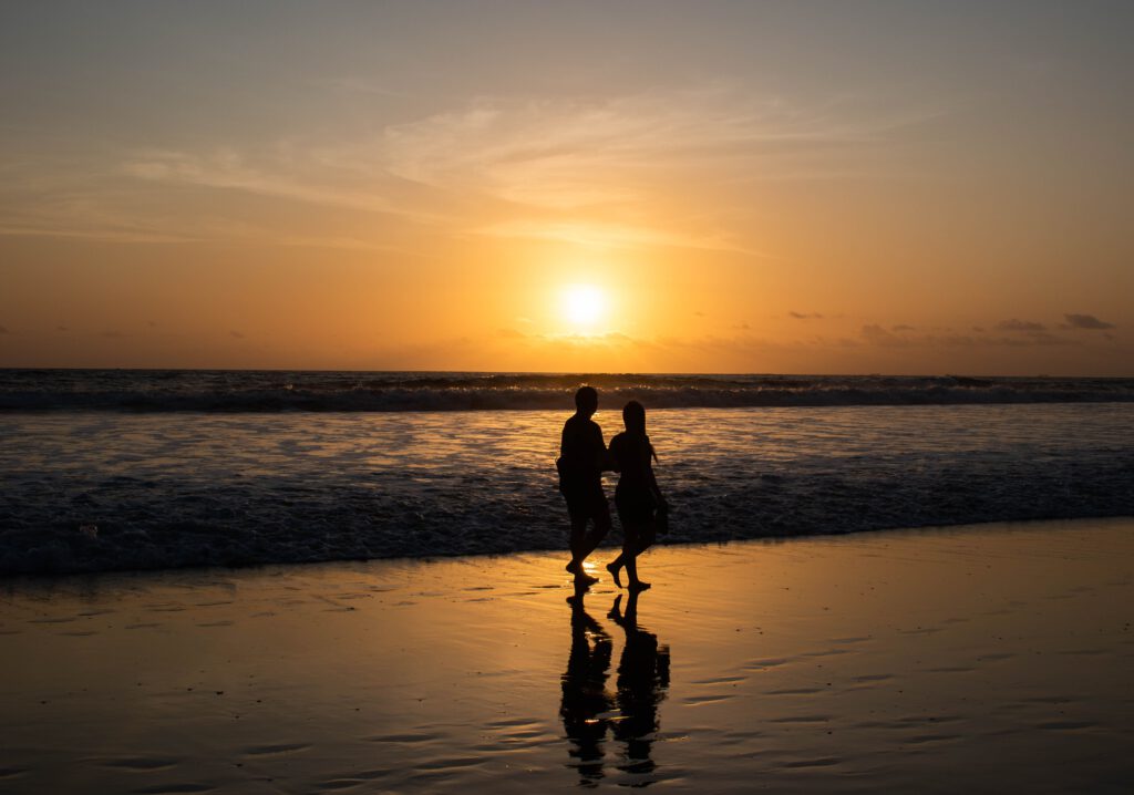 Ein Pärchen (m/w) spaziert am Strand. Die Wellen rauschen heran. Die untergehende Sonne taucht die letzten Wolken in ein orange-rotes Licht.